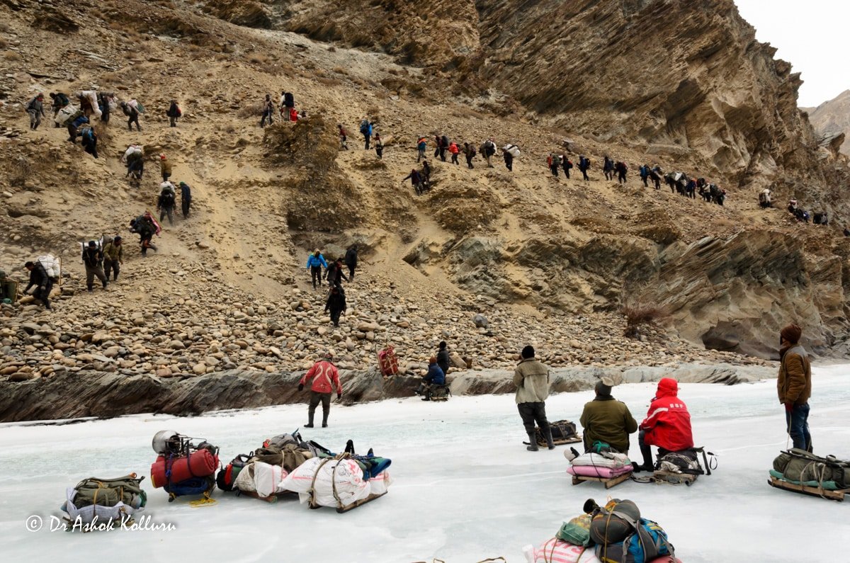 Trekkers climbing a rocky path beside a frozen river during the Chadar Trek in Ladakh