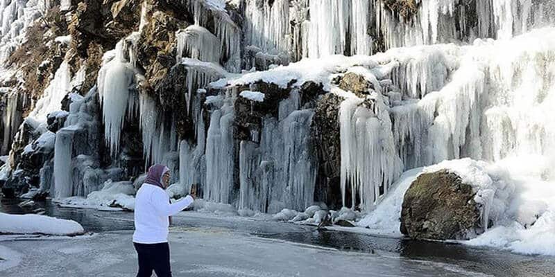 Person walking near a frozen drung waterfall with hanging icicles in a snowy winter landscape
