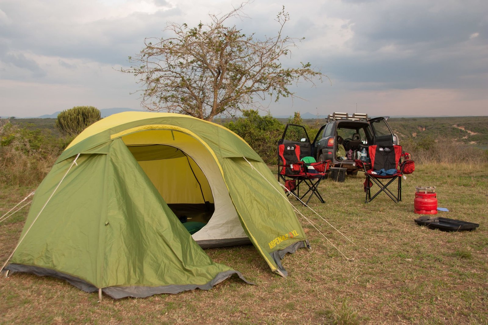 Green camping tent set up near a car with chairs and supplies in the open countryside