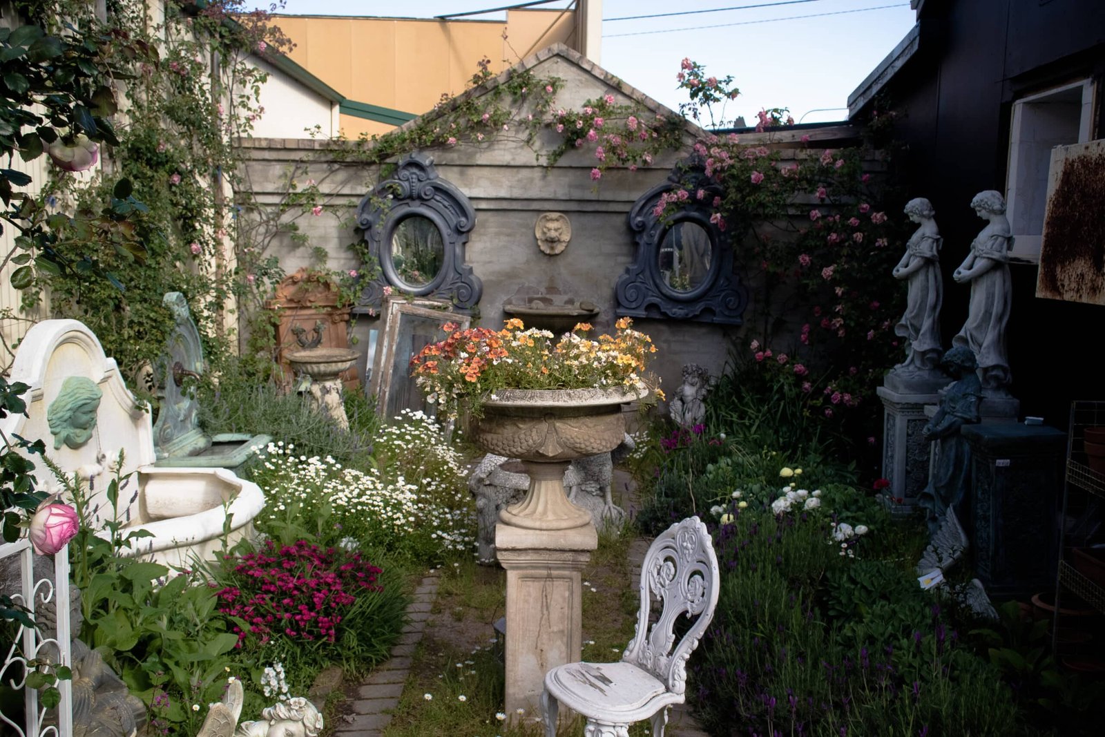 A small decorative garden with flowers, vintage chairs, and stone statues