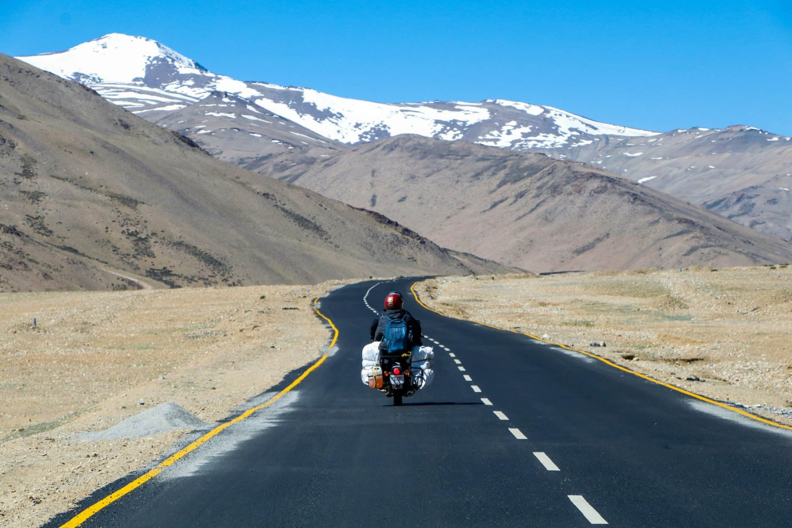 Biker riding on a winding mountain road toward snow-covered peaks in Ladakh