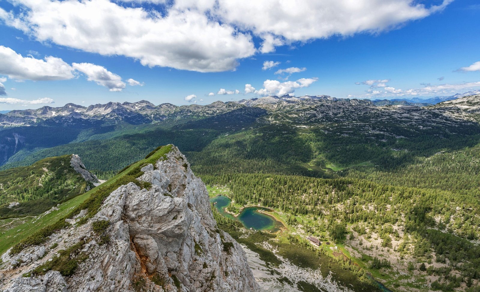 Mountain view with rocky peaks, green forest, and blue lakes under a partly cloudy sky
