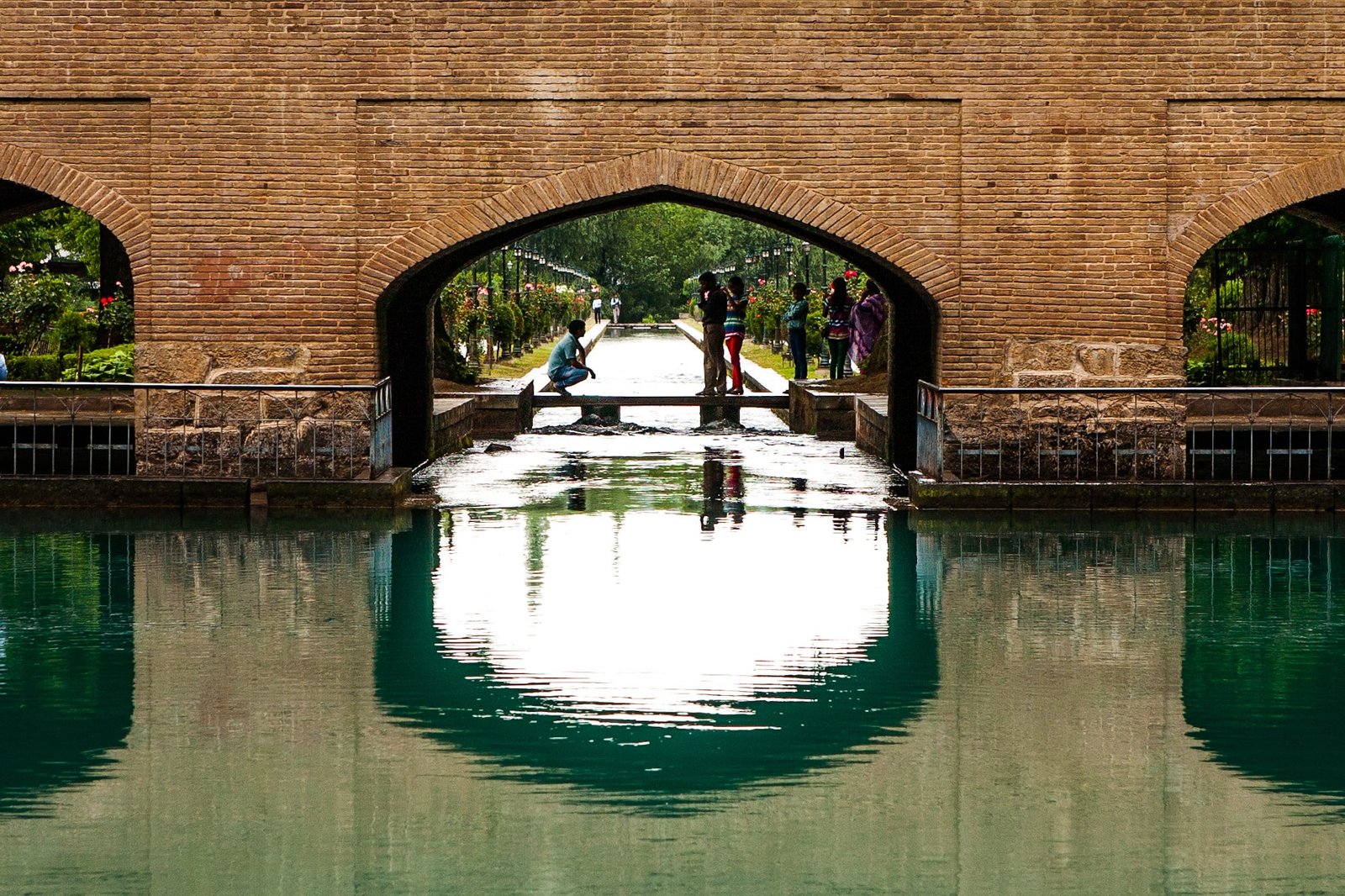 People by a water canal under an arched brick structure in a garden