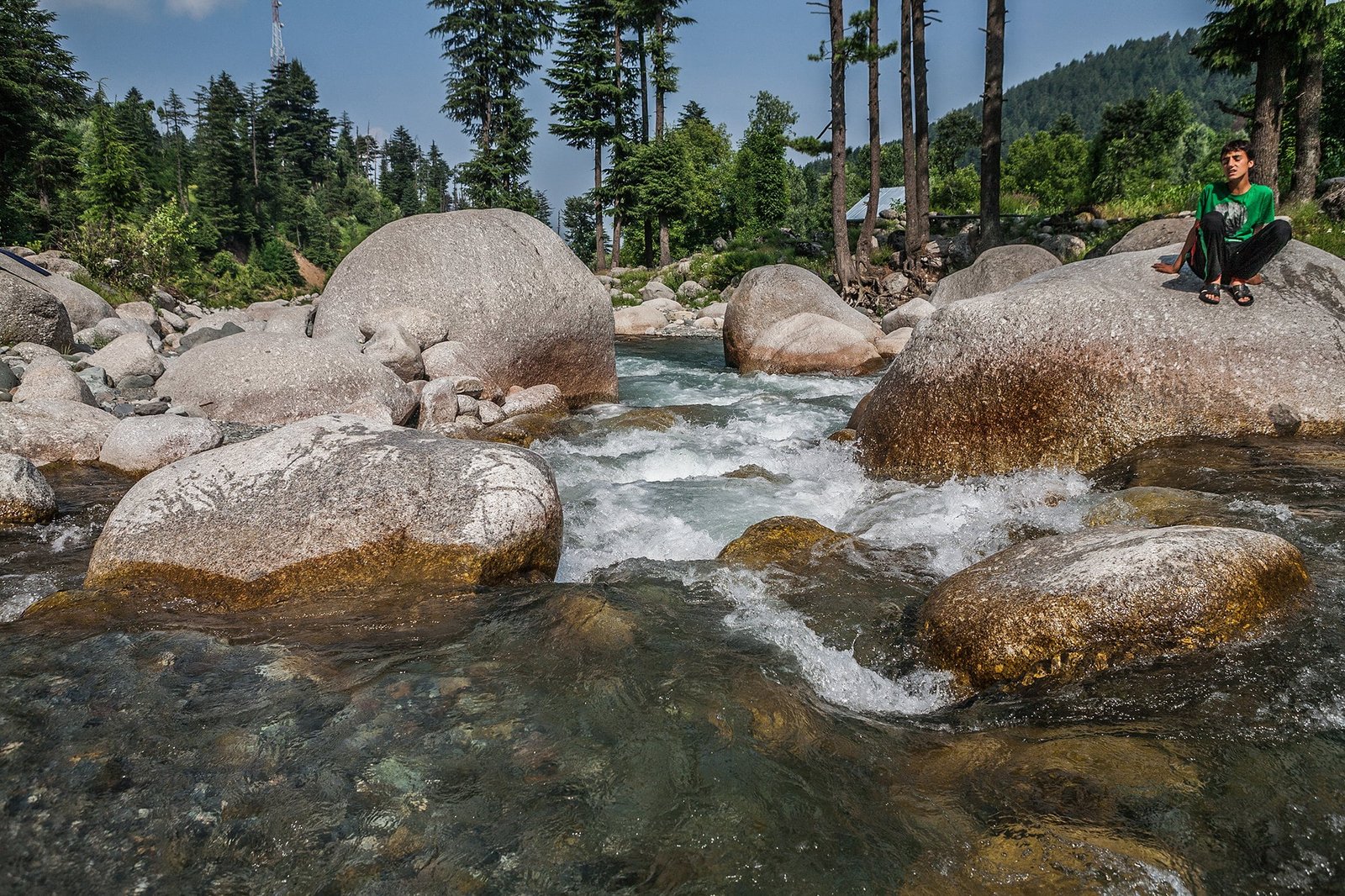 Boy sitting on a large boulder by a fast-flowing stream in a forested mountain area in Kashmir