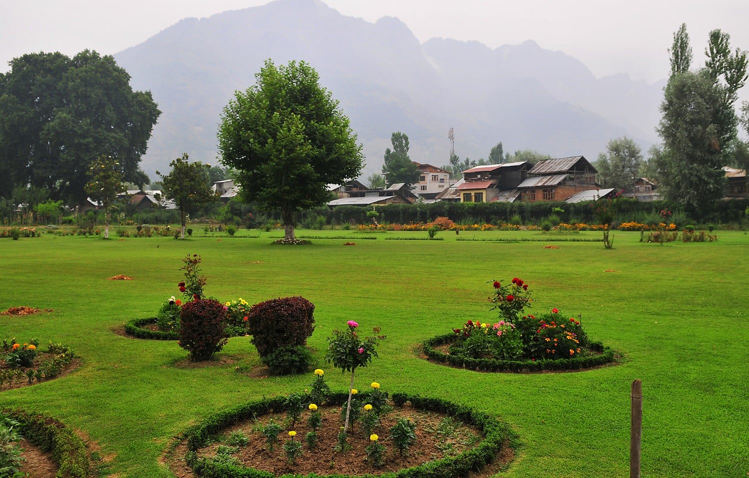 green garden with flower beds, trees, and village houses in the background