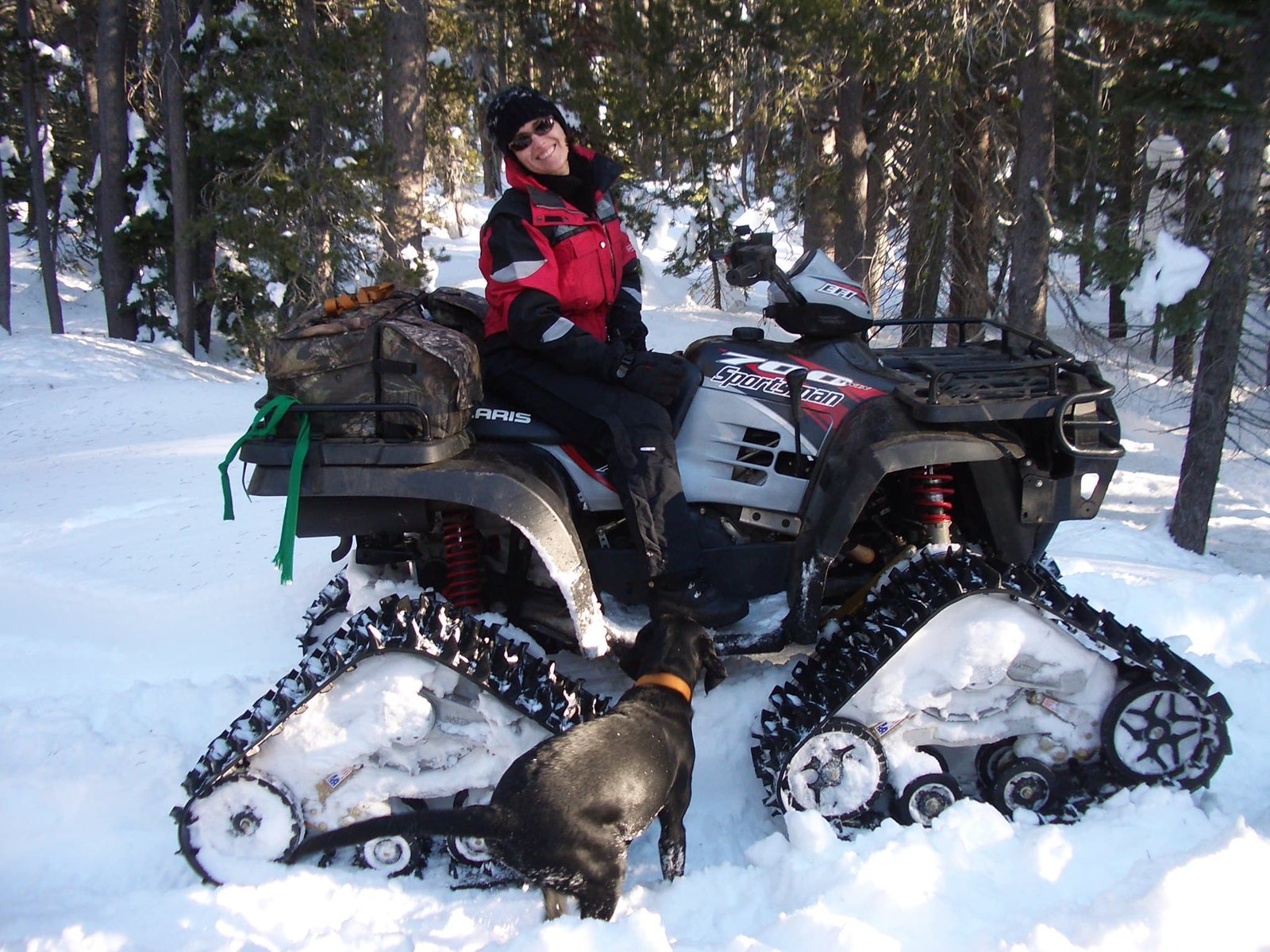 A person sits on a snow-tracked ATV in a snowy forest with a black dog nearby