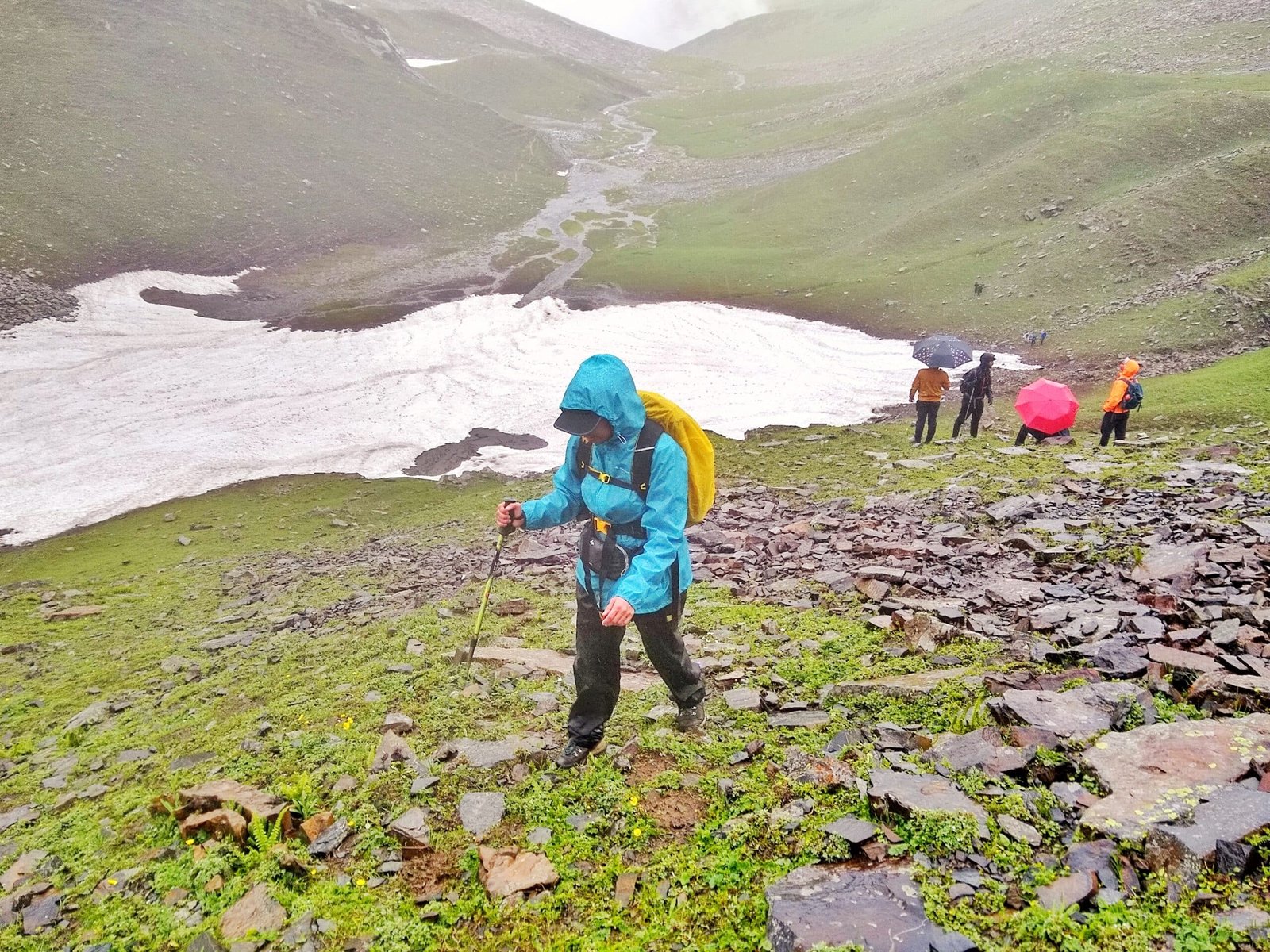 A trekker climbs a grassy and rocky slope with a trekking pole near a melting snow patch