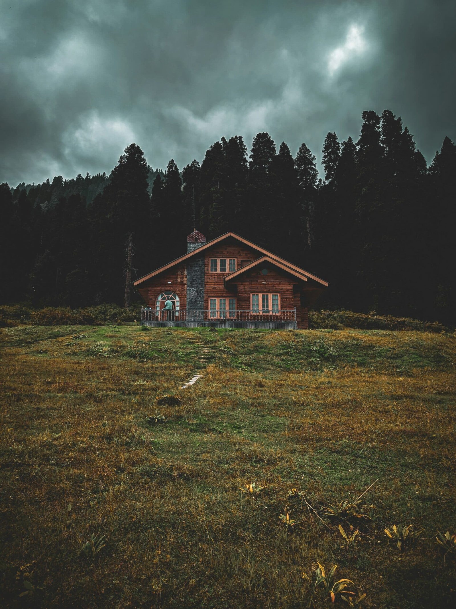 A rustic wooden cabin with a stone chimney sits on a grassy clearing, surrounded by dark pine forest under a dramatic, cloudy sky in doodhpathri