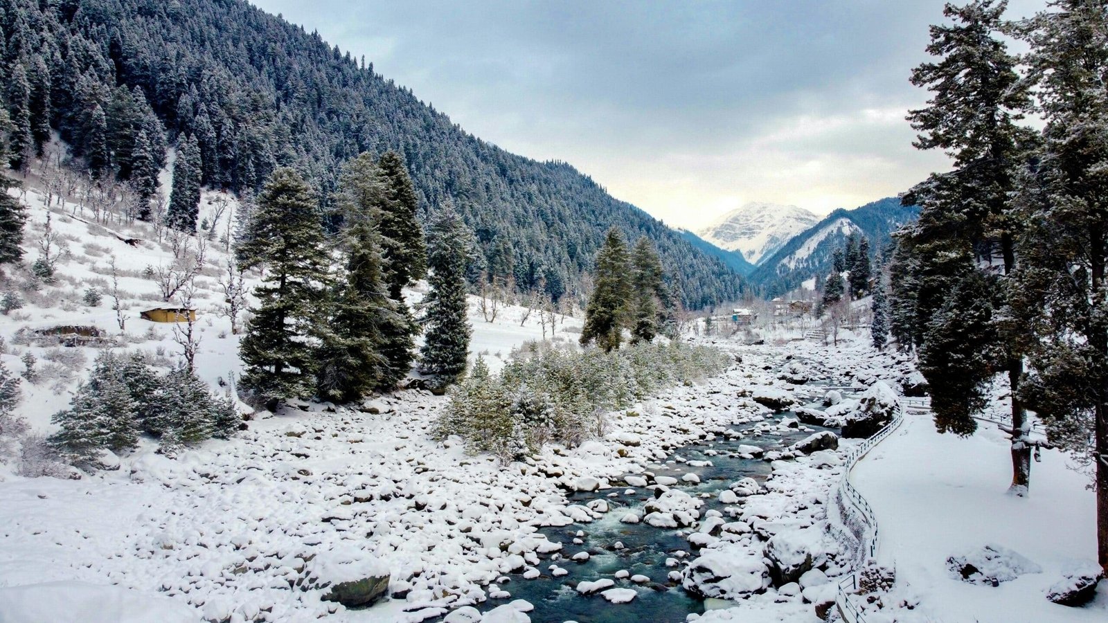 Snow-covered valley with a flowing river, pine forests, and distant snowy mountains in Pahalgam in december