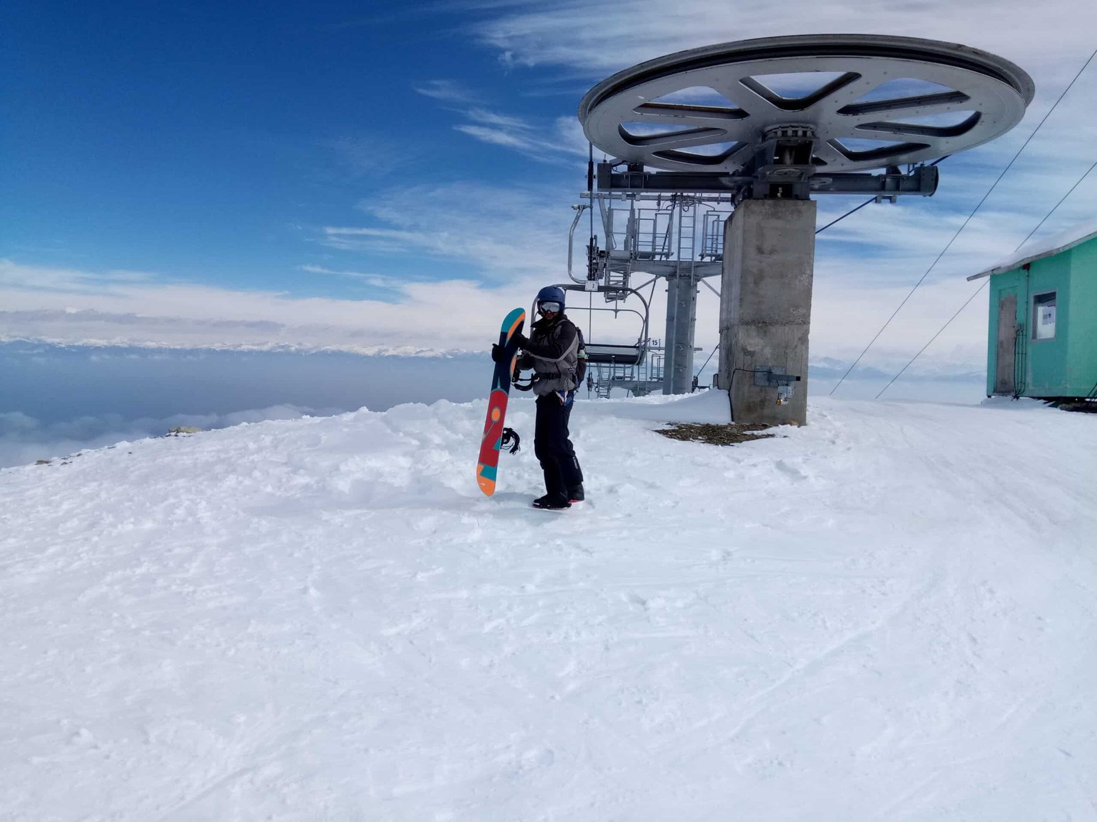 A snowboarder stands beside a ski lift at a snowy mountain summit