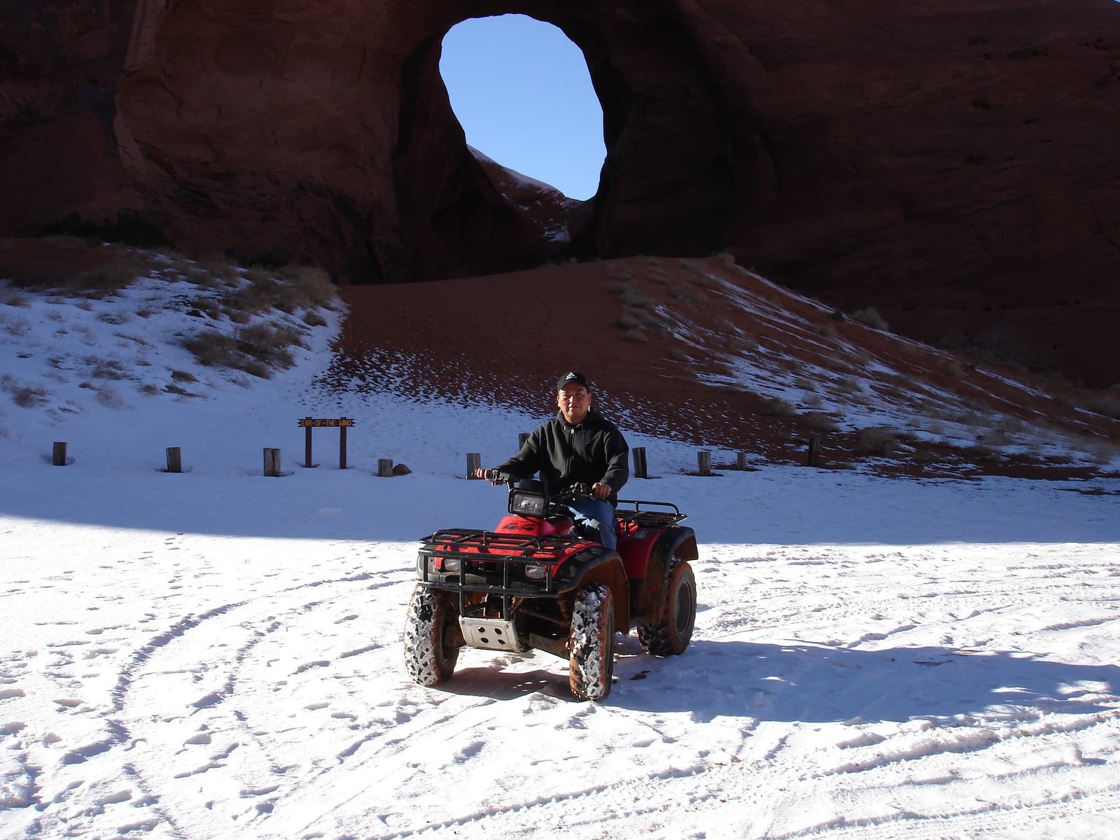 Man riding an ATV on snow at Sinthan Top with a large natural rock arch in the background.