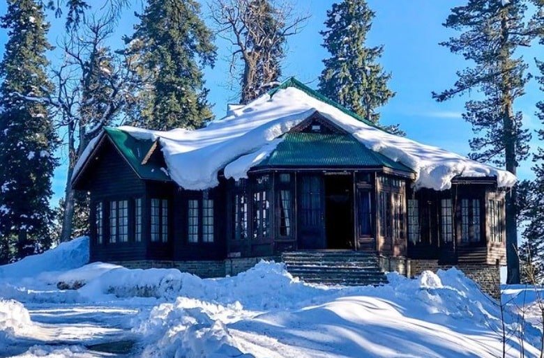 Snow-covered Maharaja Palace in Gulmarg surrounded by tall pine trees under a clear blue sky in winter
