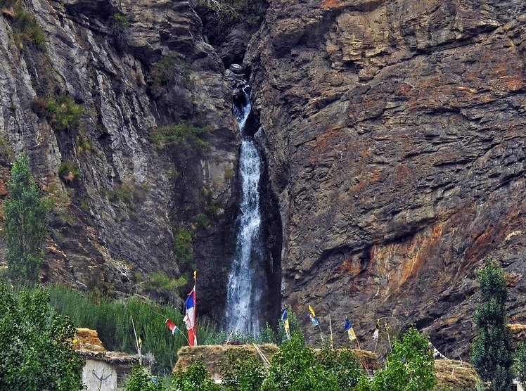 Turtuk Waterfall flowing through rocky cliffs with prayer flags and greenery below