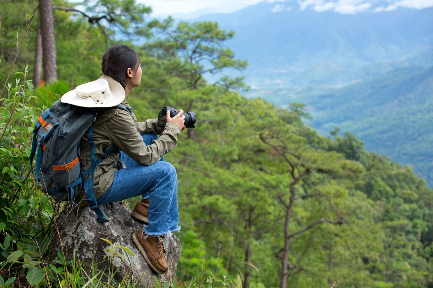 A woman with a backpack and camera sits on a rock overlooking a forested mountain valley
