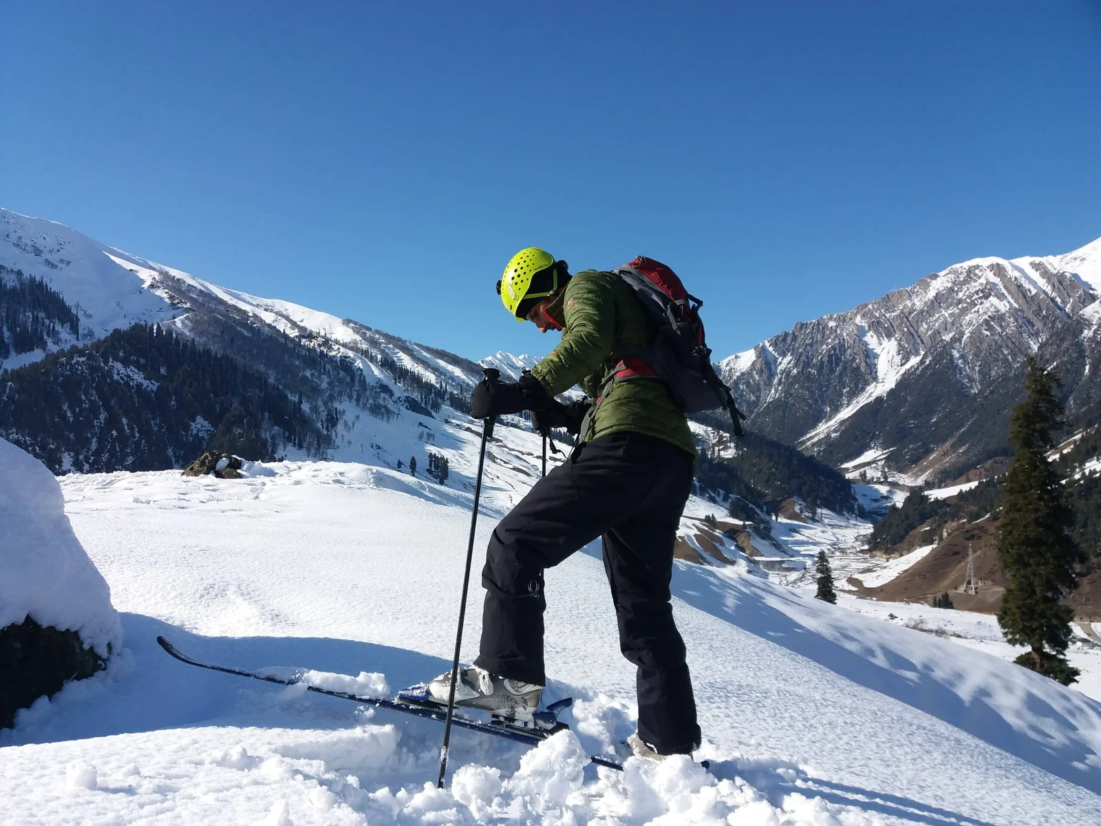 A skier in a green jacket and yellow helmet prepares to descend a snowy slope in Gulmarg